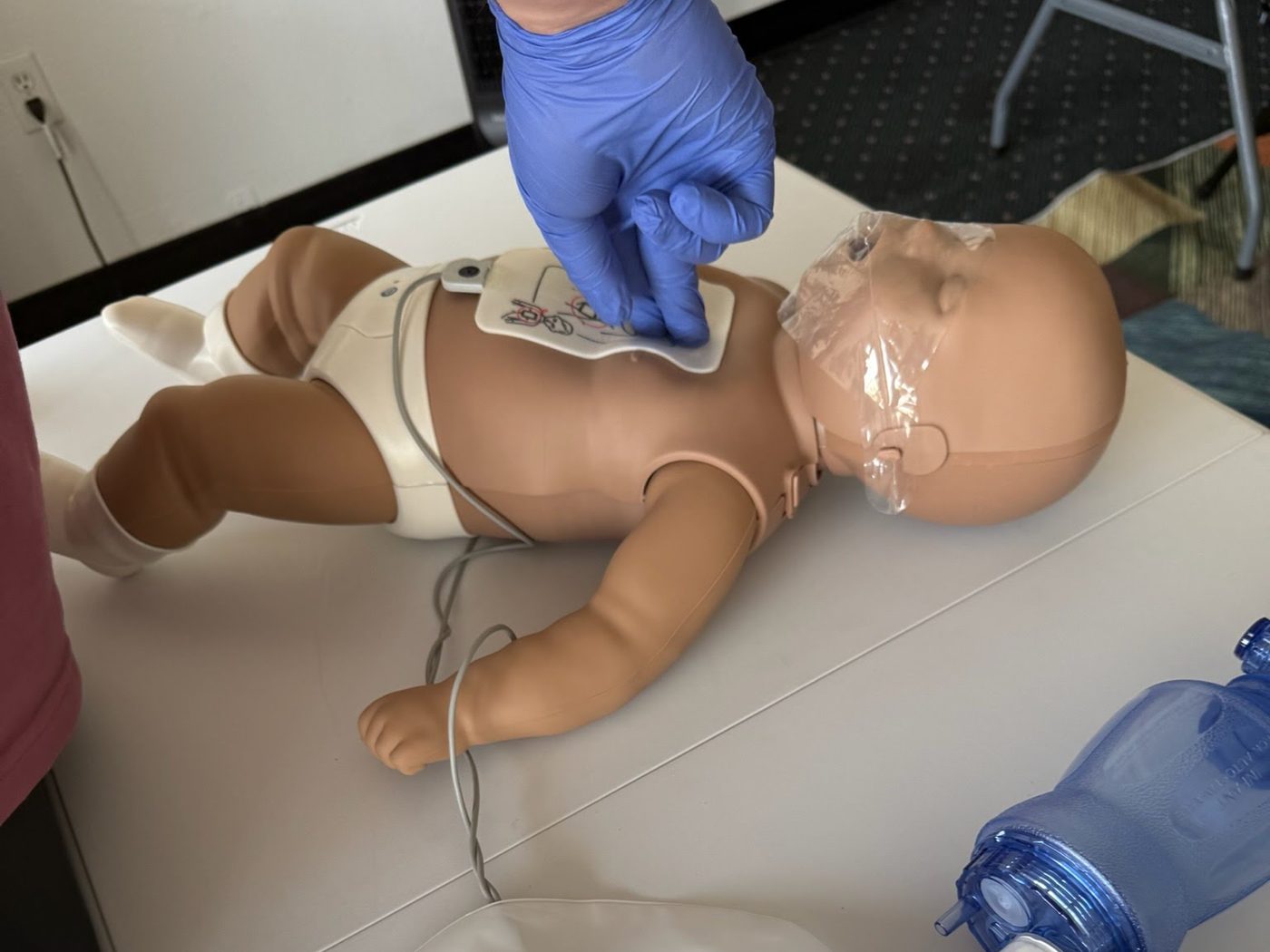 Gloved hands practicing infant CPR on an infant manikin with an AED trainer and bag-valve mask